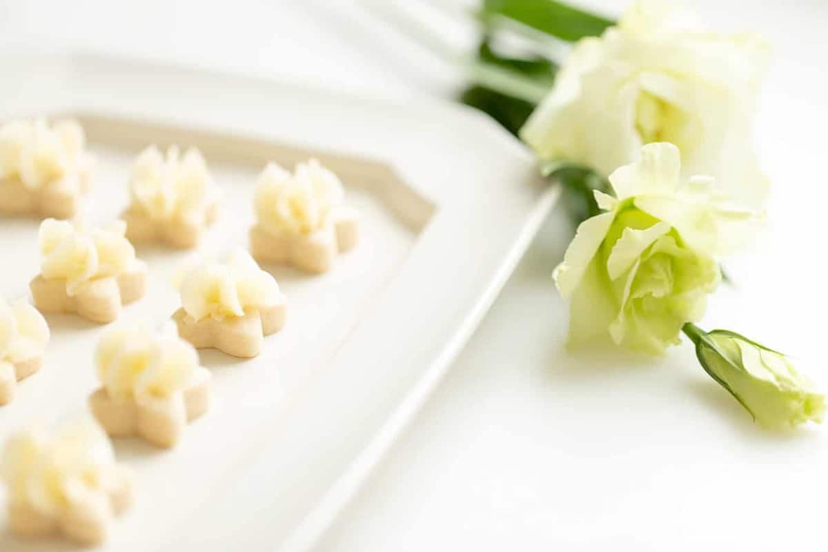 Small flower shaped cookies, frosted with lemon icing on a white platter. Flowers are to the side. 