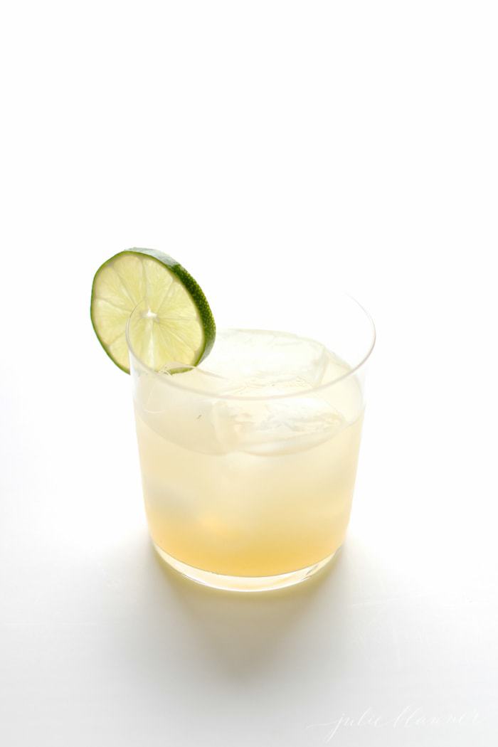A white surface with a single tumbler of a margarita with ice, lime garnish.