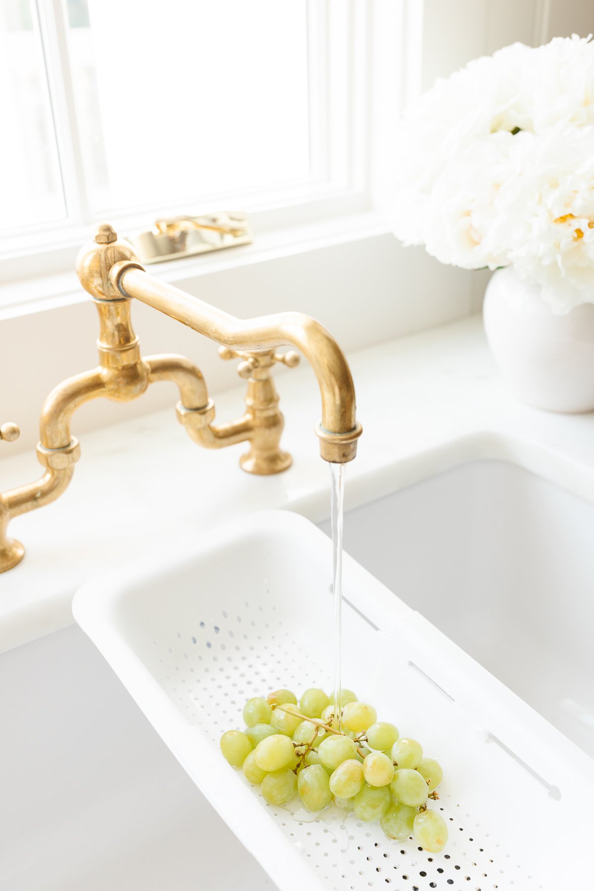 A brass faucet running over a white strainer with grapes in a white farmhouse sink.