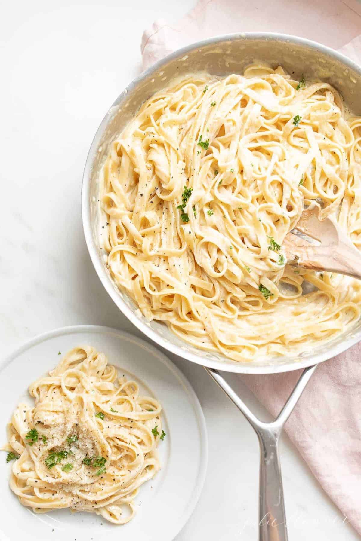 White surface with a metal pan full of homemade fettuccine alfredo, single plate of pasta to the side.