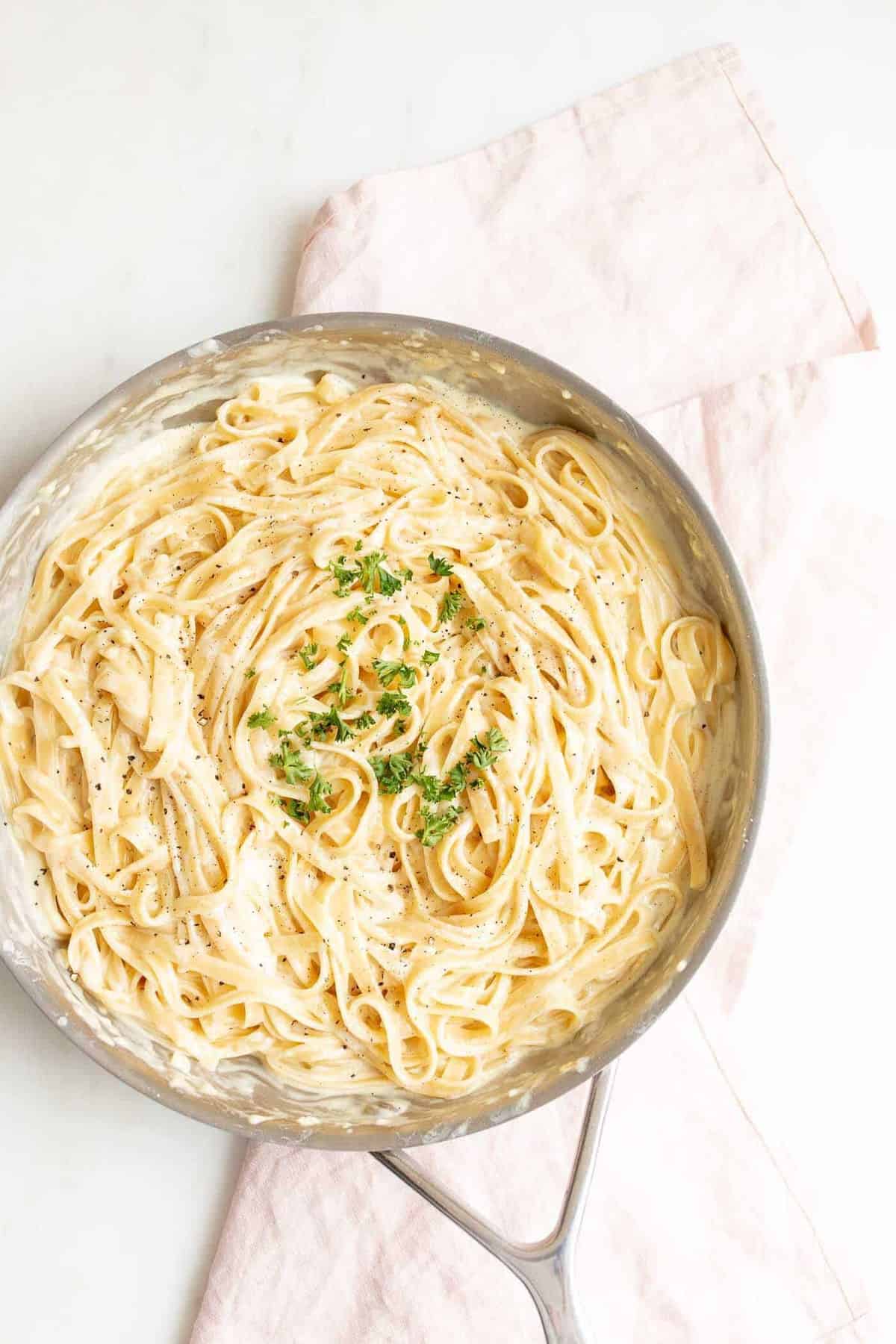 Fettuccine Alfredo in a metal pan on a white surface with a pink linen napkin to the side.