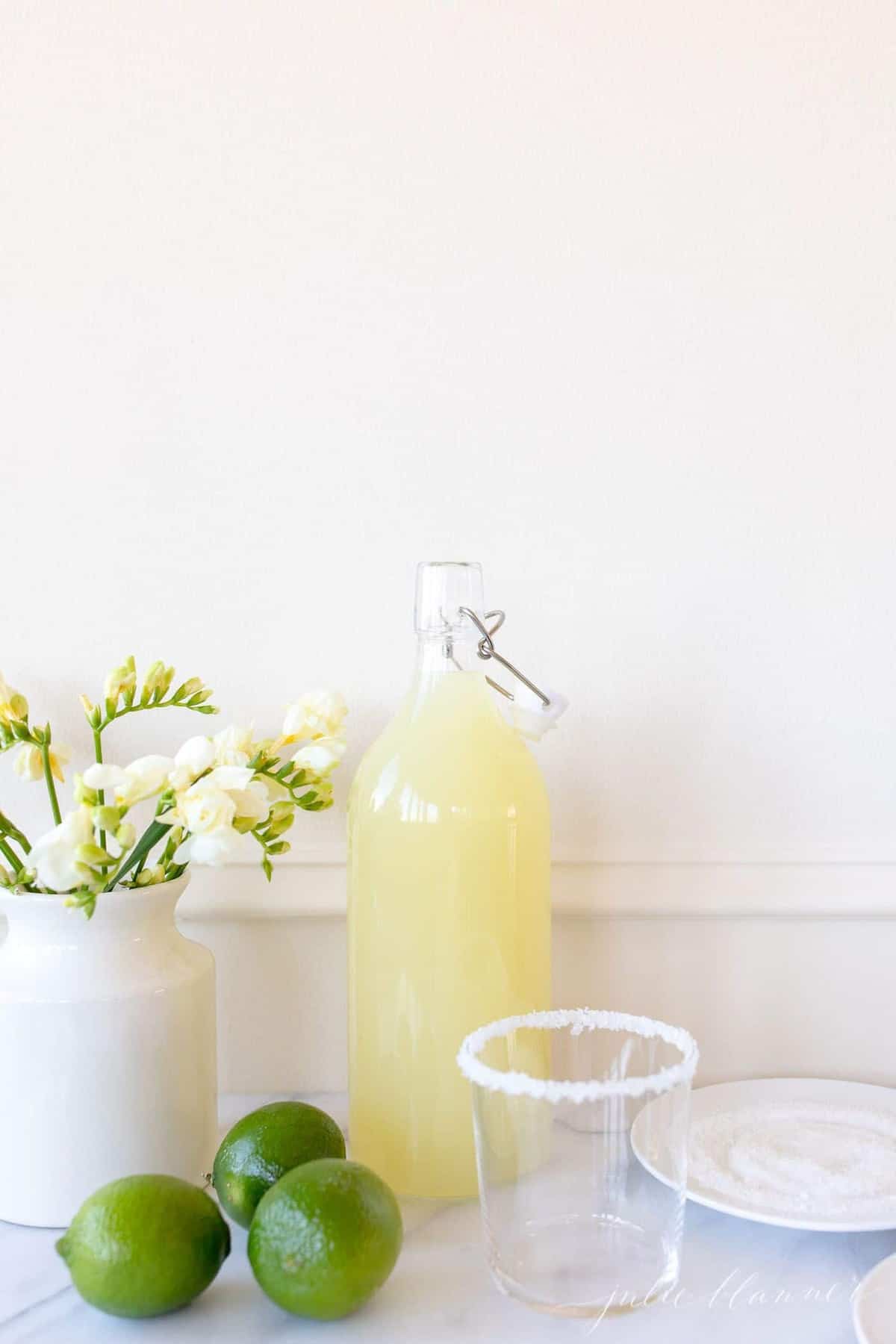 Homemade margarita mix in a glass carafe; a vase full of yellow flowers sits to the side. 