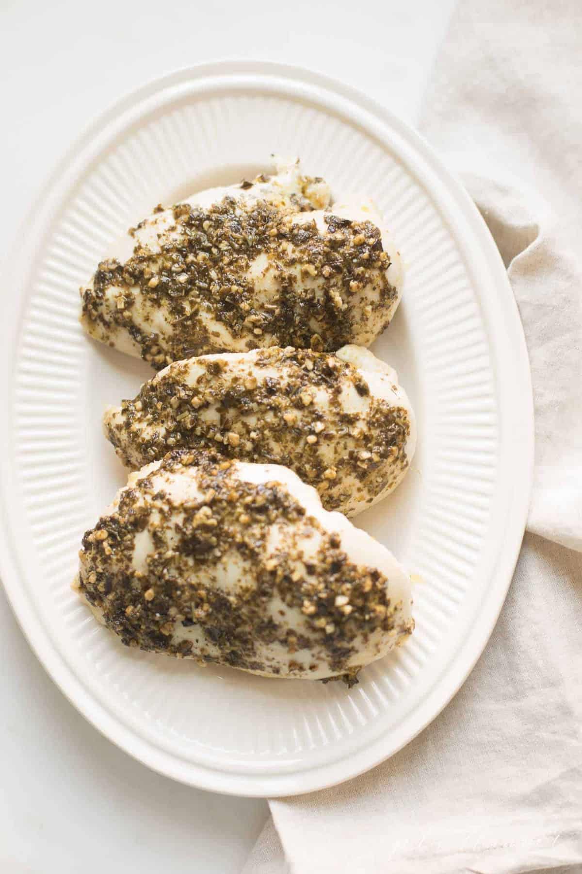 An oval platter with three chicken breasts covered in homemade basil pesto sauce.