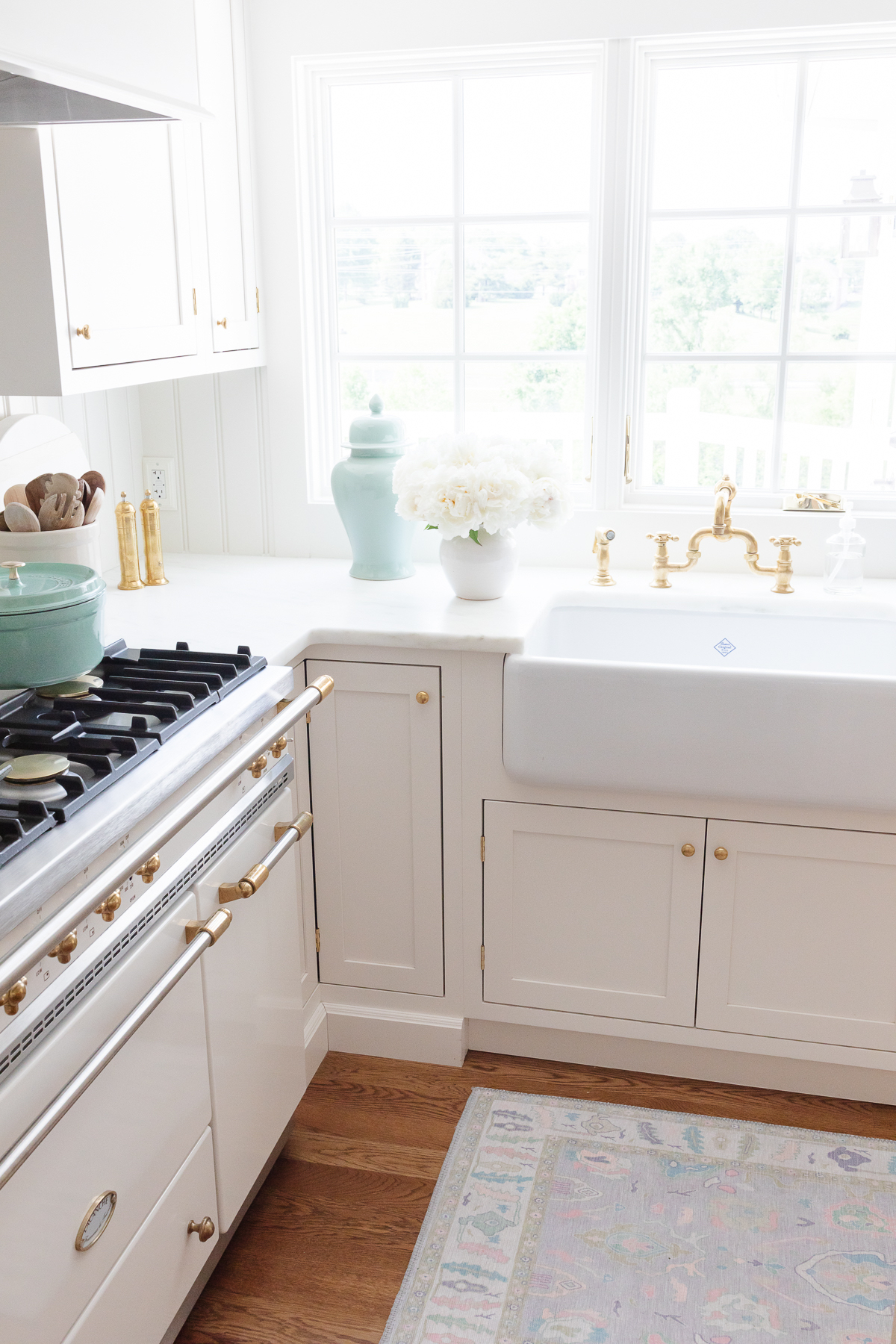 A bright kitchen with white cabinets, a farmhouse sink, gold fixtures, a stove with brass accents, and a window. Decorative items include a vase with flowers and a pastel blue ceramic jar.