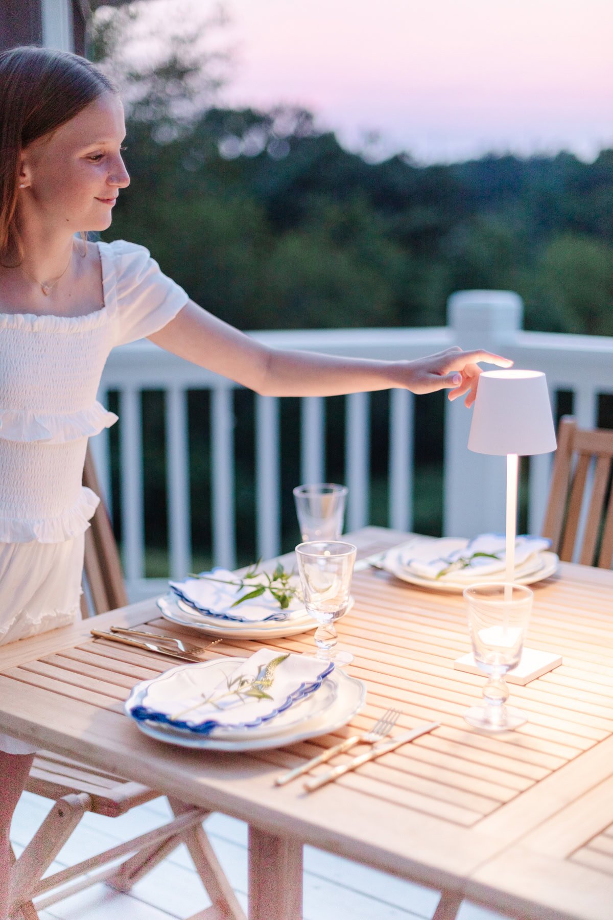 A girl in a white dress adjusts a lamp on a wooden outdoor dining table set with plates, glasses, and cutlery at sunset.