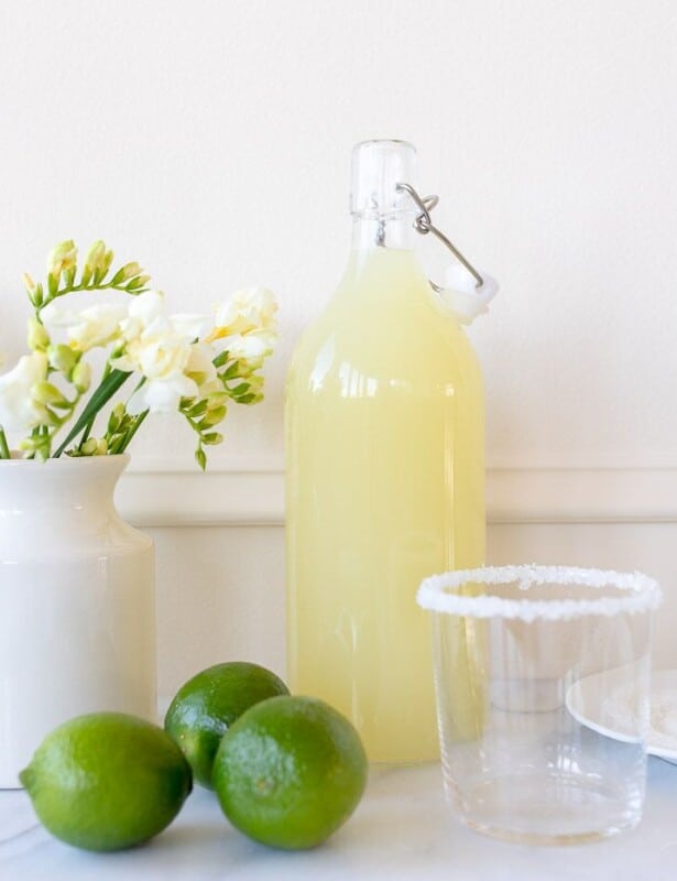 White kitchen background, glass carafe full of homemade margarita mix, limes to the side.