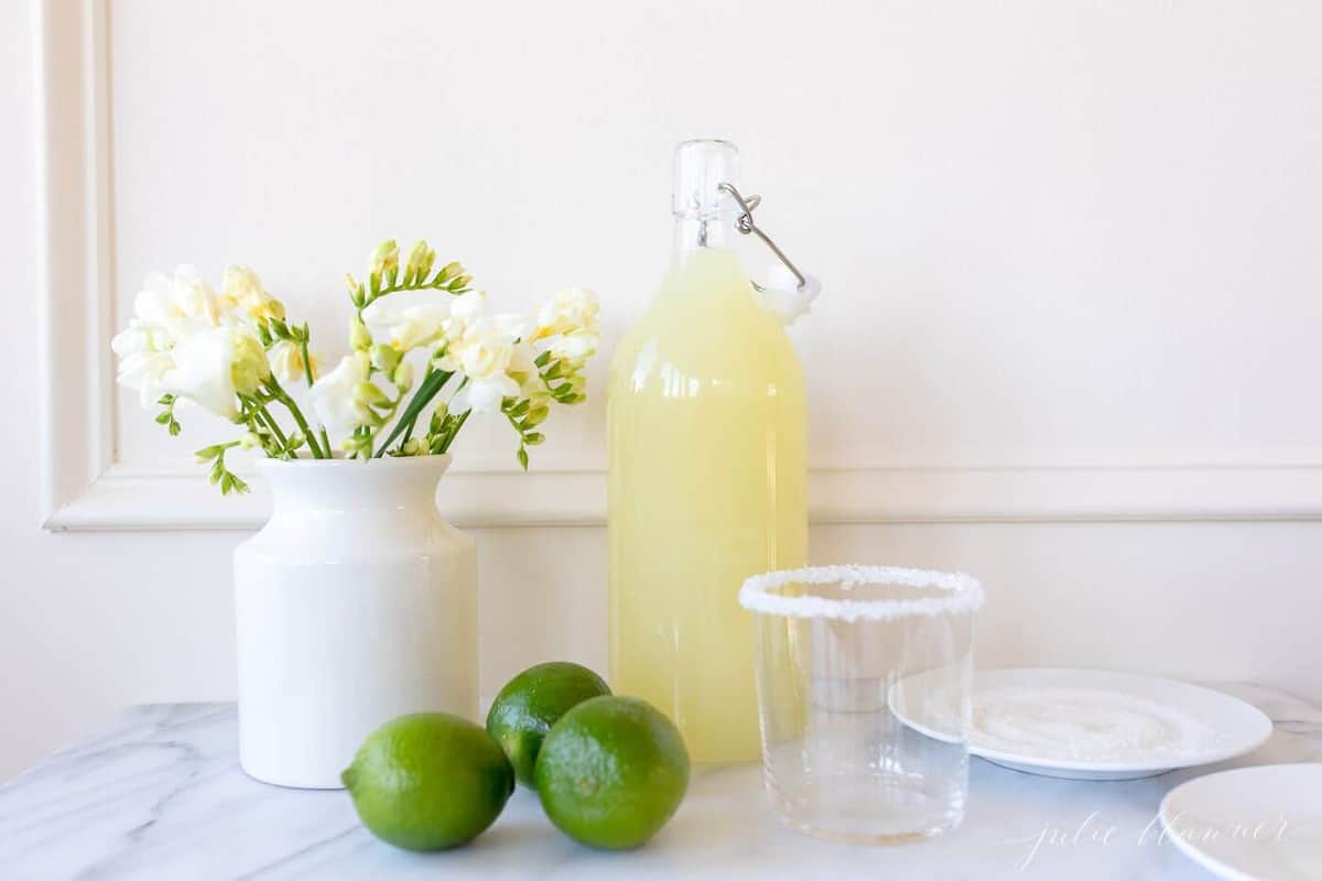 White kitchen background, glass carafe full of homemade margarita mix, limes to the side.