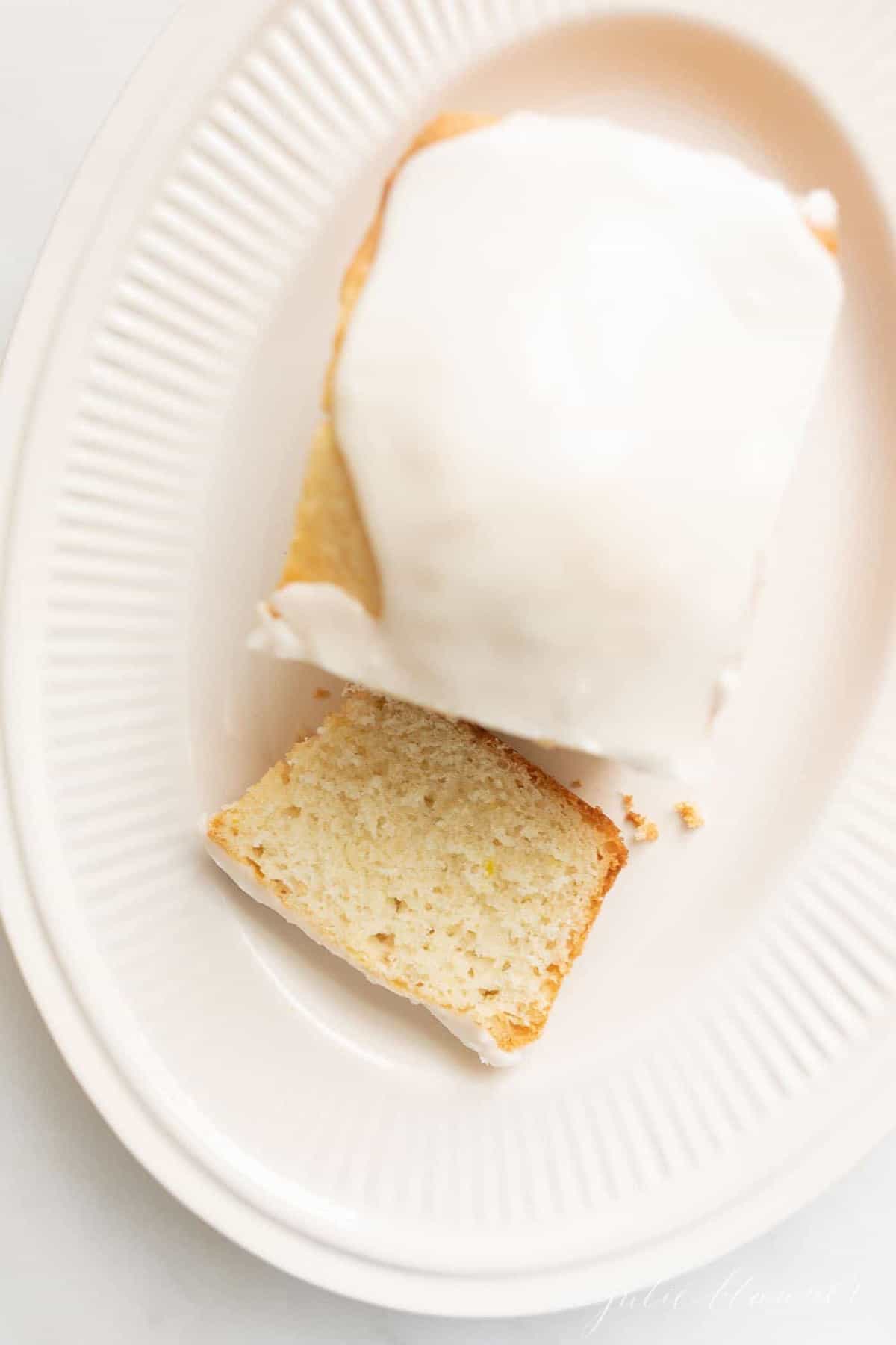 A loaf of iced lemon quick bread, one slice cut on an ivory platter.
