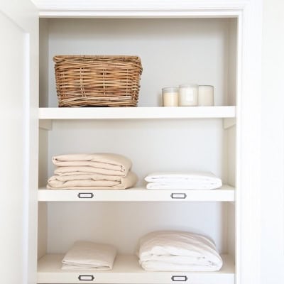 A linen closet with three shelves holding a wicker basket, candles in glass containers, neatly folded white and beige linens, and drawer organizers keeping everything tidy.