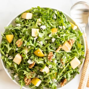 A bowl of arugula salad with pear slices, walnuts, and crumbled blue cheese. Matching serving utensils with woven handles are placed beside the bowl on a white surface.