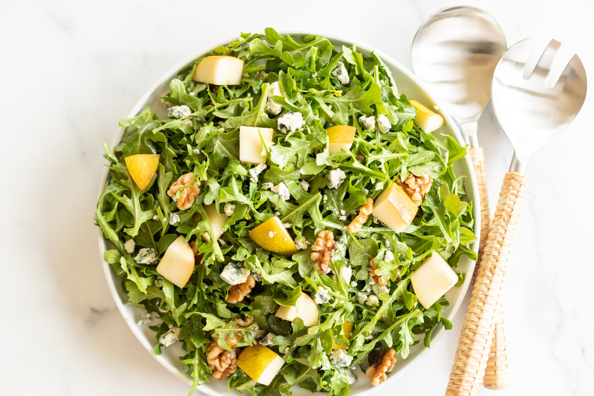 A bowl of arugula salad with pear slices, walnuts, and crumbled blue cheese. Matching serving utensils with woven handles are placed beside the bowl on a white surface.