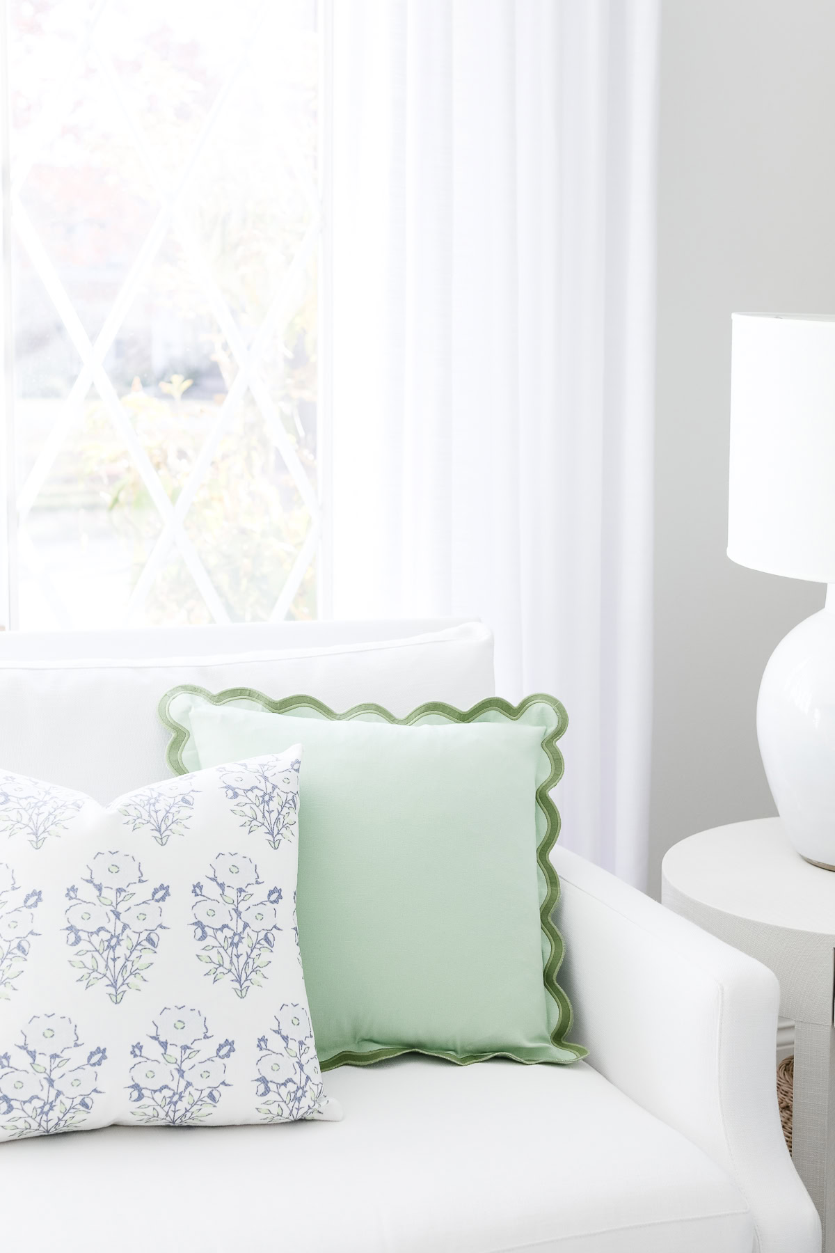 A white couch with two decorative pillows—a green one with scalloped edges and a white one with a blue floral pattern—next to a round white side table with a white lamp.