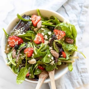 A fresh spring salad with berries, crumbled cheese and nuts in a bowl with wooden serving utensils.