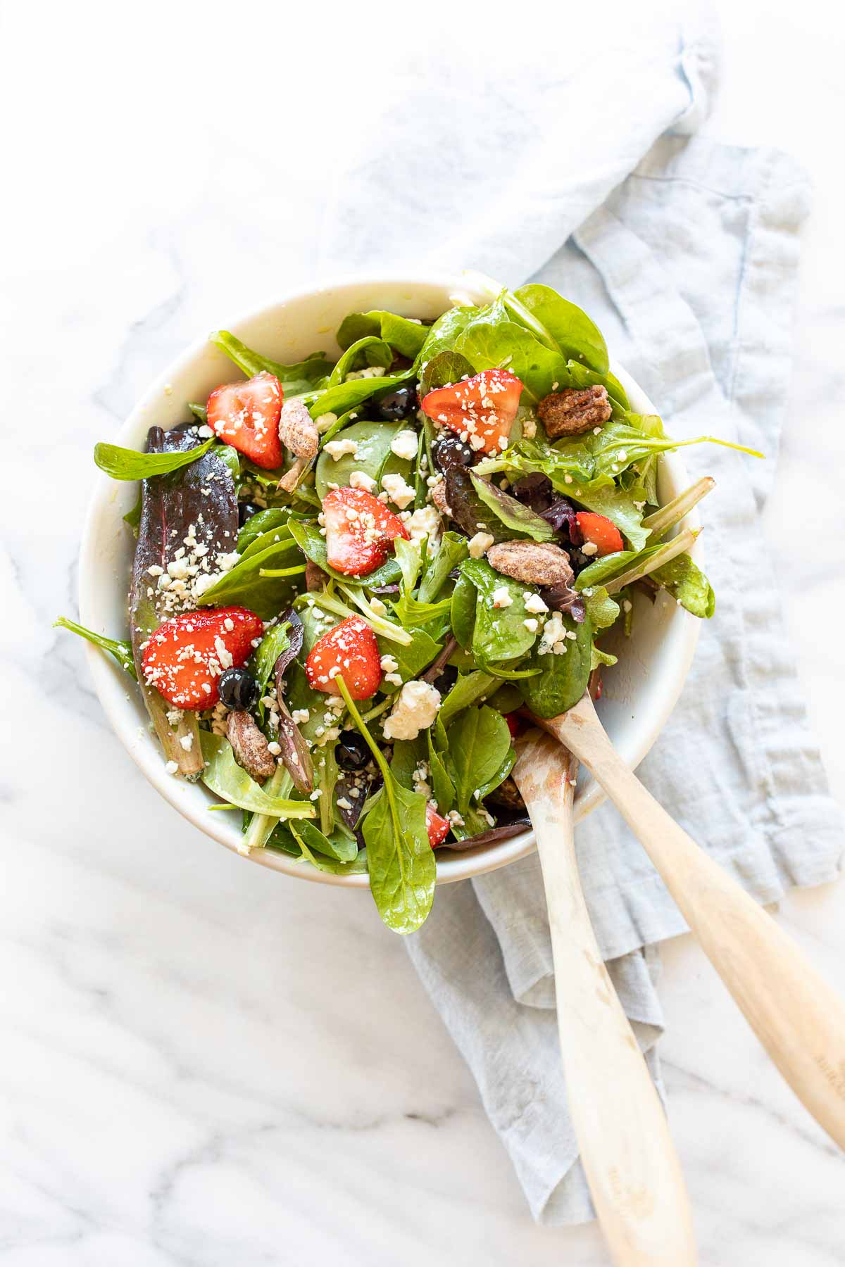 A fresh spring salad with berries, crumbled cheese and nuts in a bowl with wooden serving utensils.