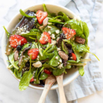 A fresh spring salad with berries, crumbled cheese and nuts in a bowl with wooden serving utensils.