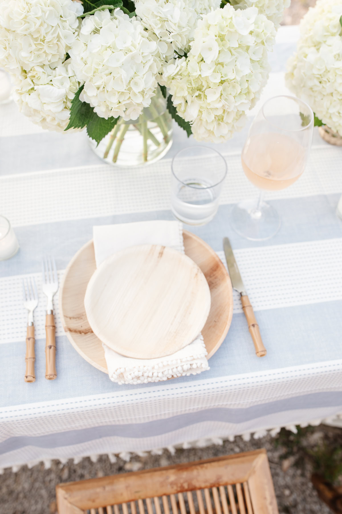 Elegant table setting with a wooden plate, silverware with bamboo handles, a glass of rosé wine, and white hydrangea flower arrangements.