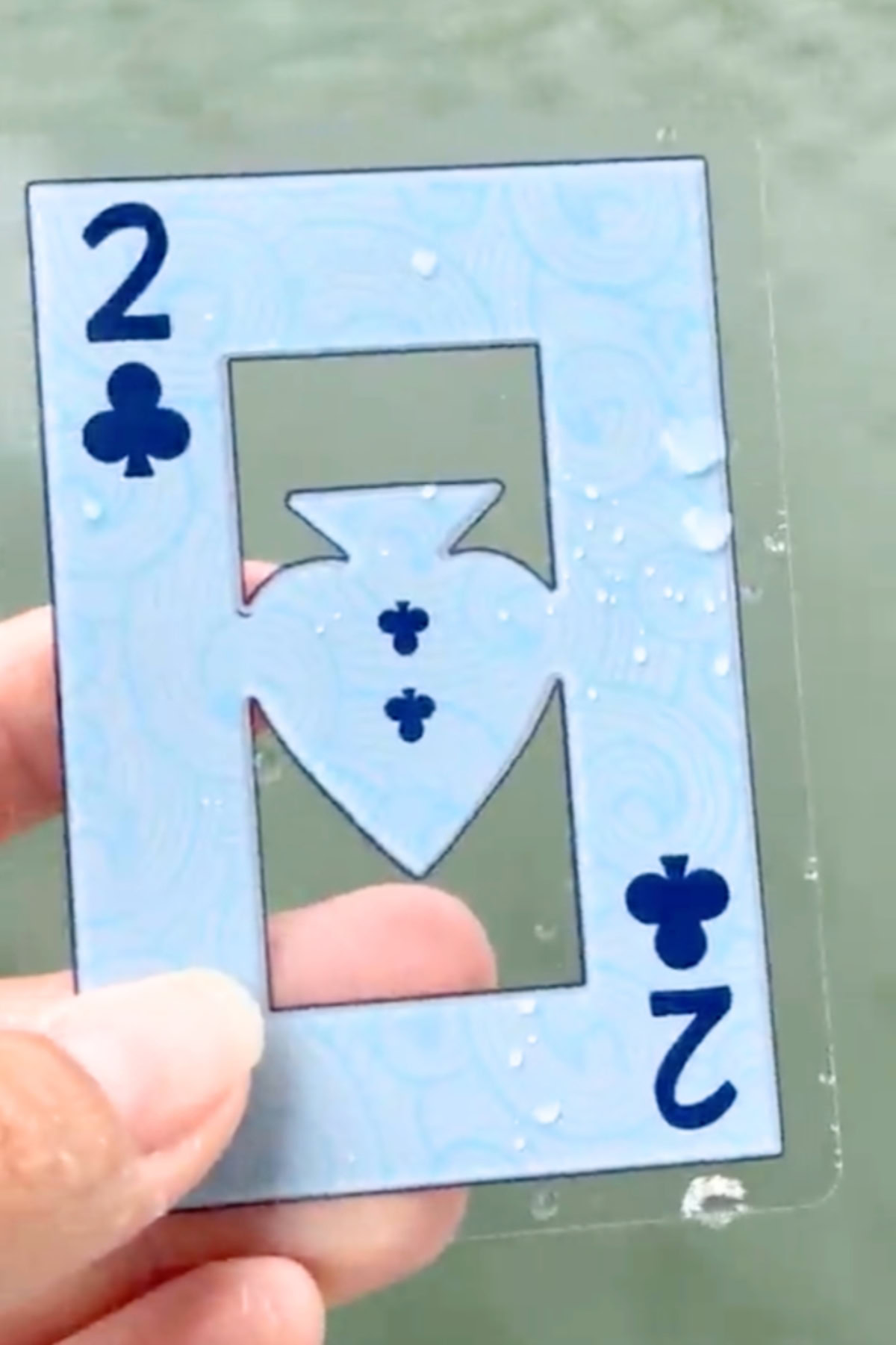 Hand holding a transparent playing card of 2 of clubs with a cutout design and water droplets on it.