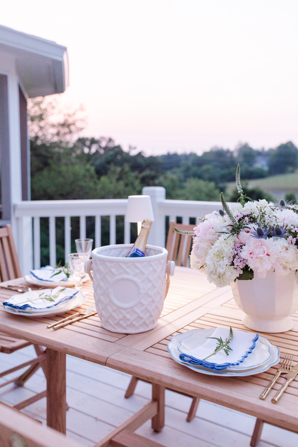 A wooden outdoor dining table set with plates, cutlery, napkins, a white ice bucket with a bottle, and a flower arrangement, overlooking a scenic view with a railing and greenery in the background.