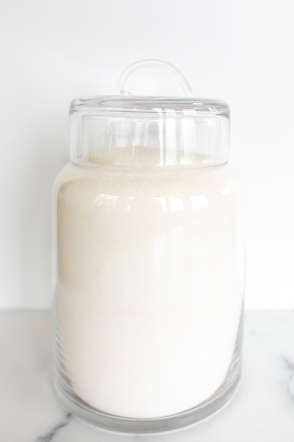 A clear glass jar filled with white sugar, sealed with a lid, sitting on a white marble surface, ideal for baking substitutions.