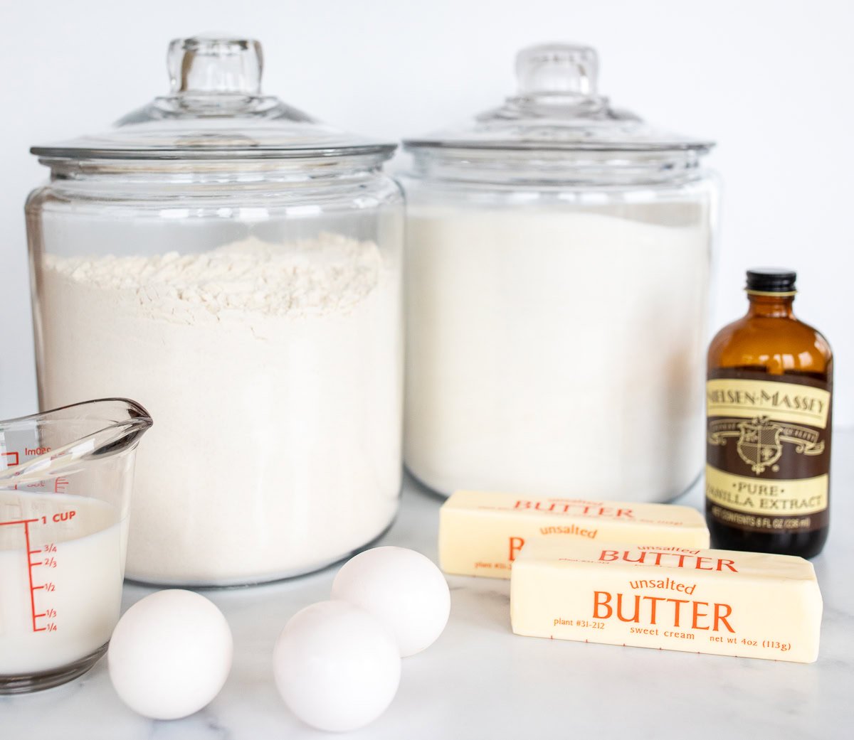 Ingredients and baking substitutions arranged on a kitchen counter, including jars of flour and sugar, eggs, butter, a glass measuring cup, and vanilla extract.