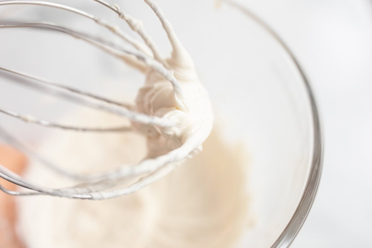 Whisk with whipped cream clinging to it, positioned above a glass bowl against a white background, perfect for exploring baking substitutions.
