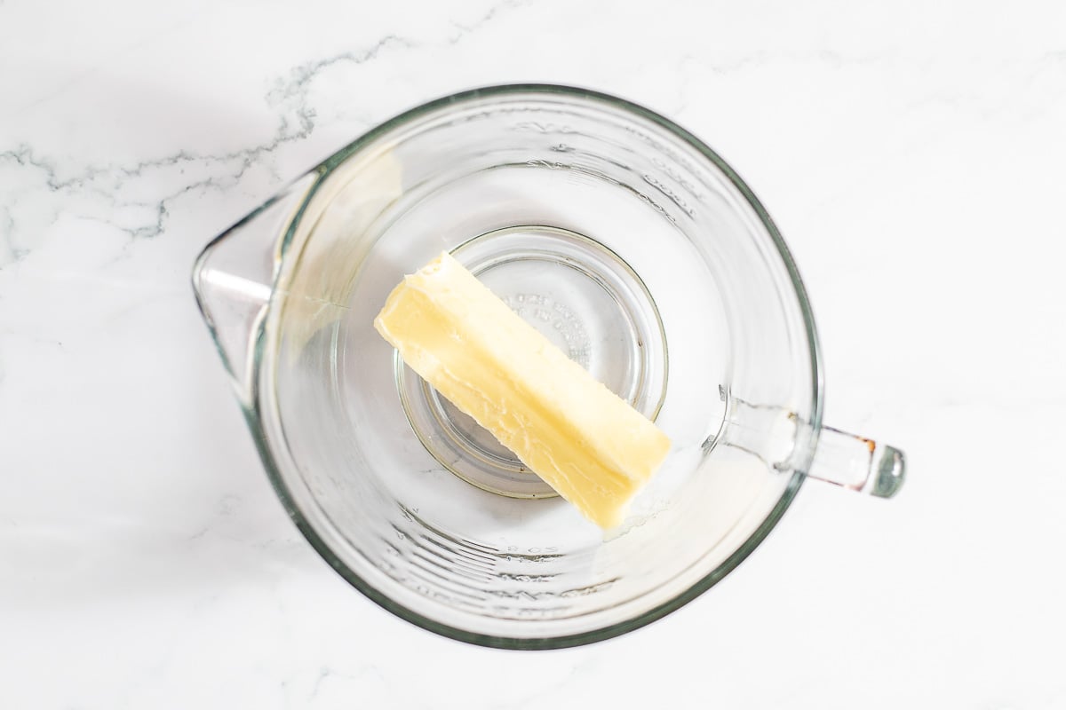A stick of butter and baking substitutions in a glass measuring cup on a marble surface.