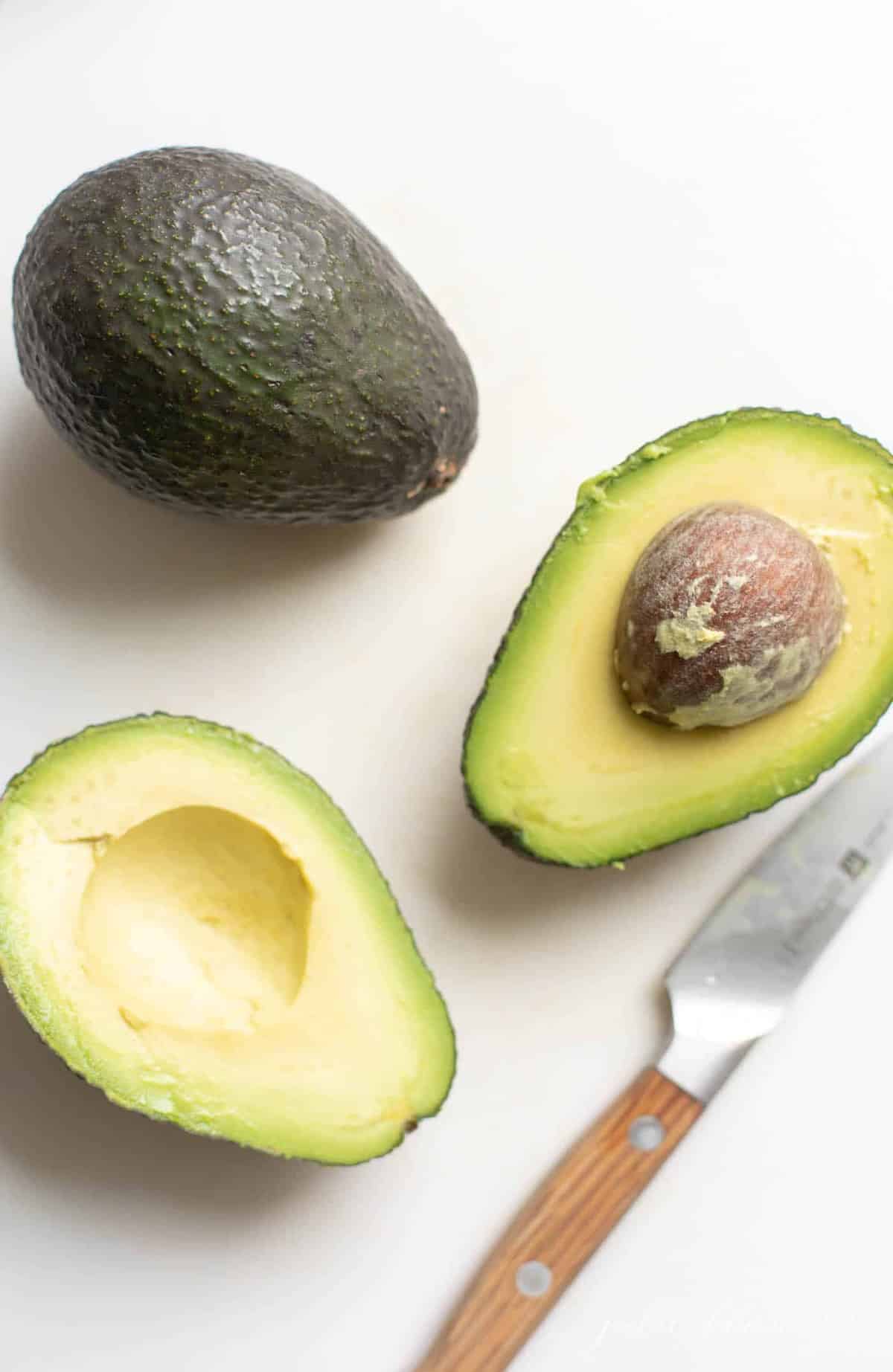 A white surface with two avocados, one sliced open with a knife to the side.