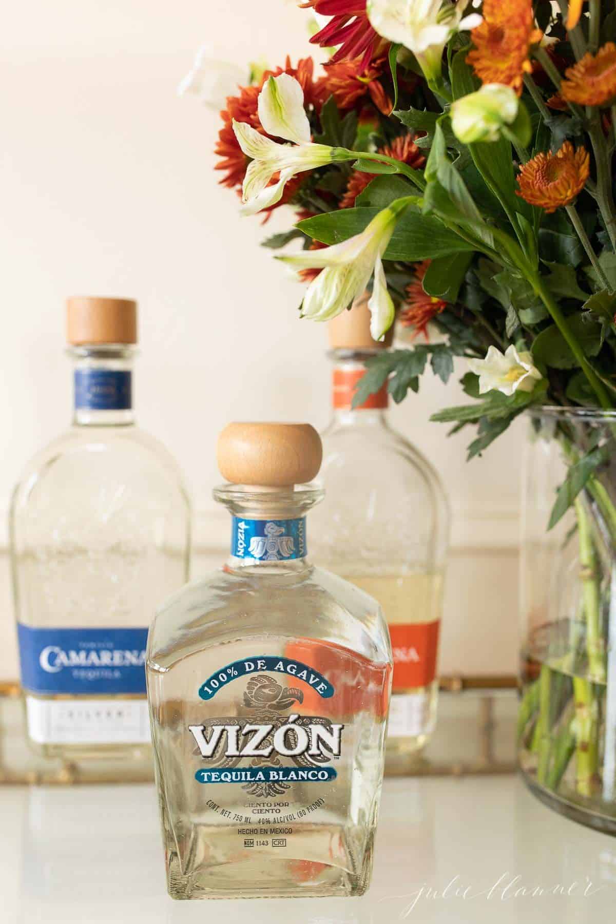 Three clear glass bottles of the best tequila for margaritas on a glass bar cart, flowers to the side.