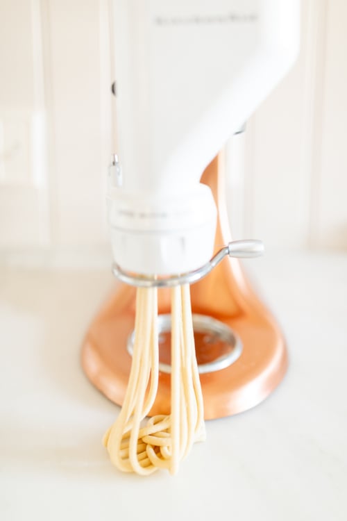 A kitchen stand mixer in white with a pasta maker attachment creating fresh bucatini noodles, set on a light marble counter.
