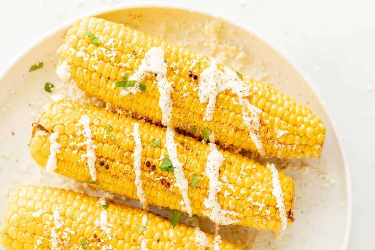 A white platter with an elote recipe of three mexican corn on the cobs, drizzled with cream sauce and cilantro.