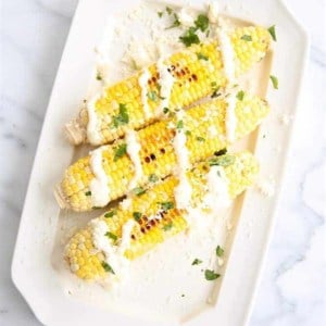 A white platter with an elote recipe of three Mexican corn cobs, drizzled with cream sauce and coriander.