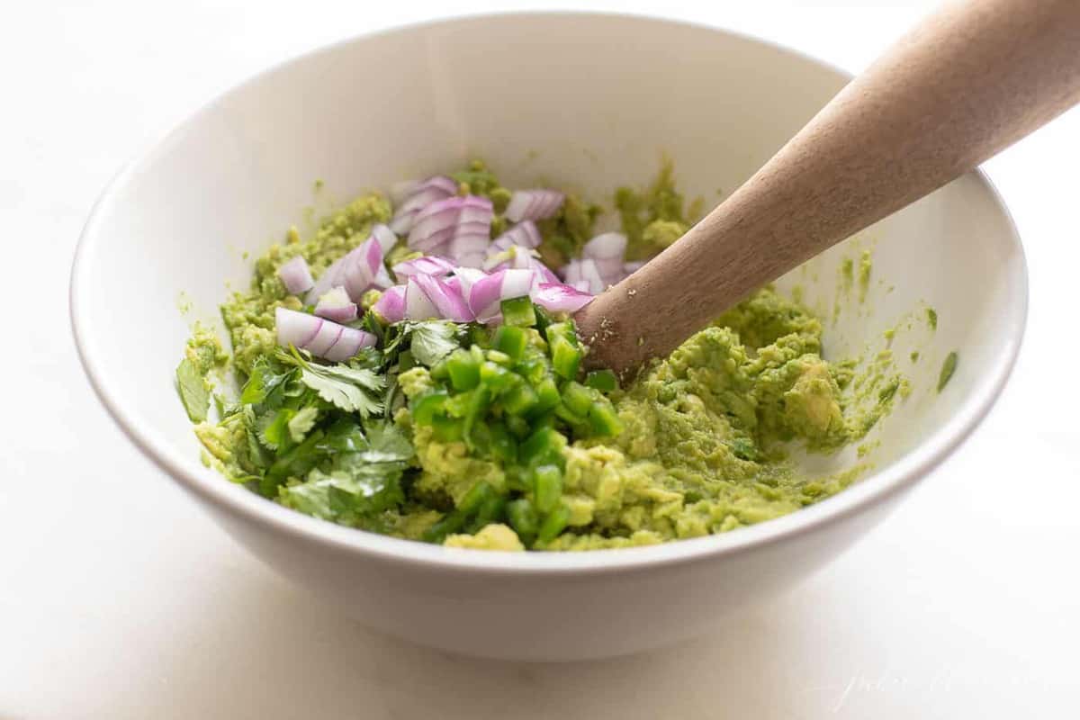 A white bowl with mashed avocados and a wooden spoon, starting a homemade guacamole recipe.