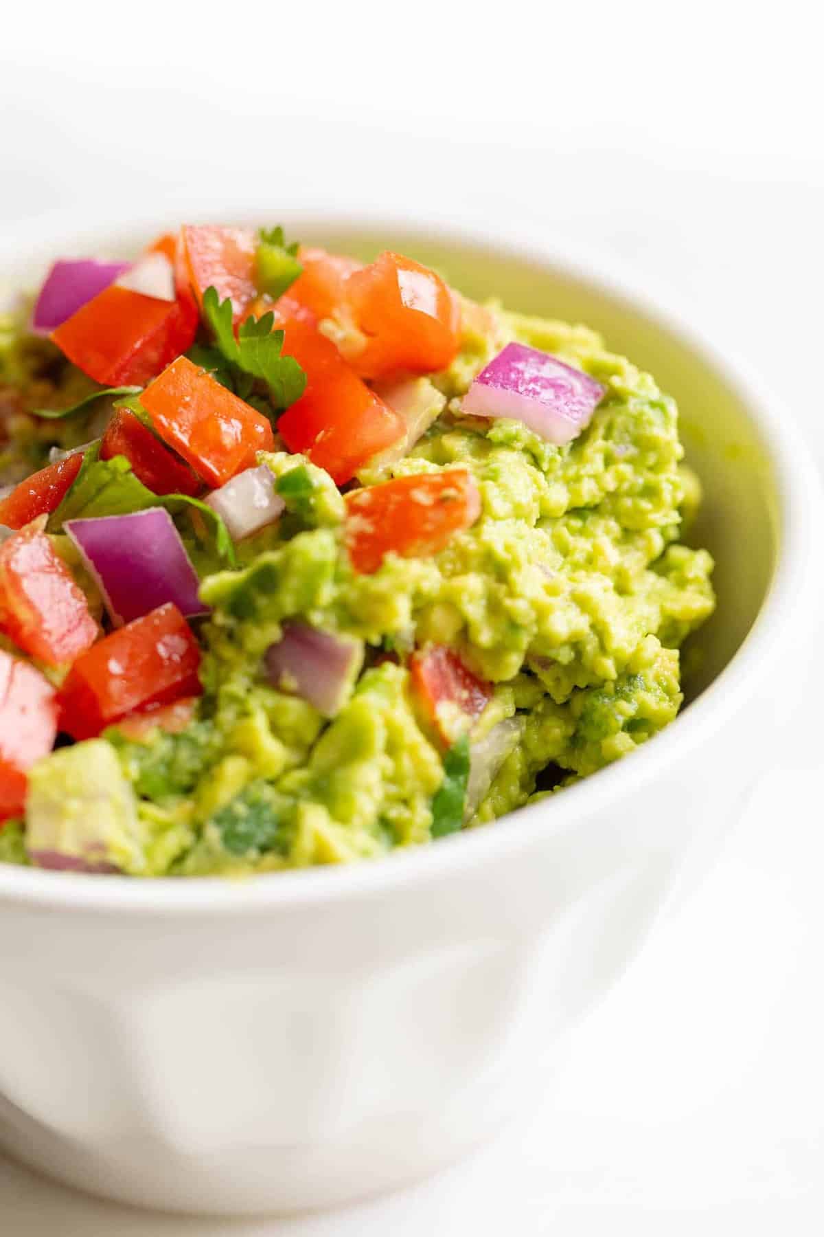 A white bowl on a white surface, filled with a homemade guacamole recipe.