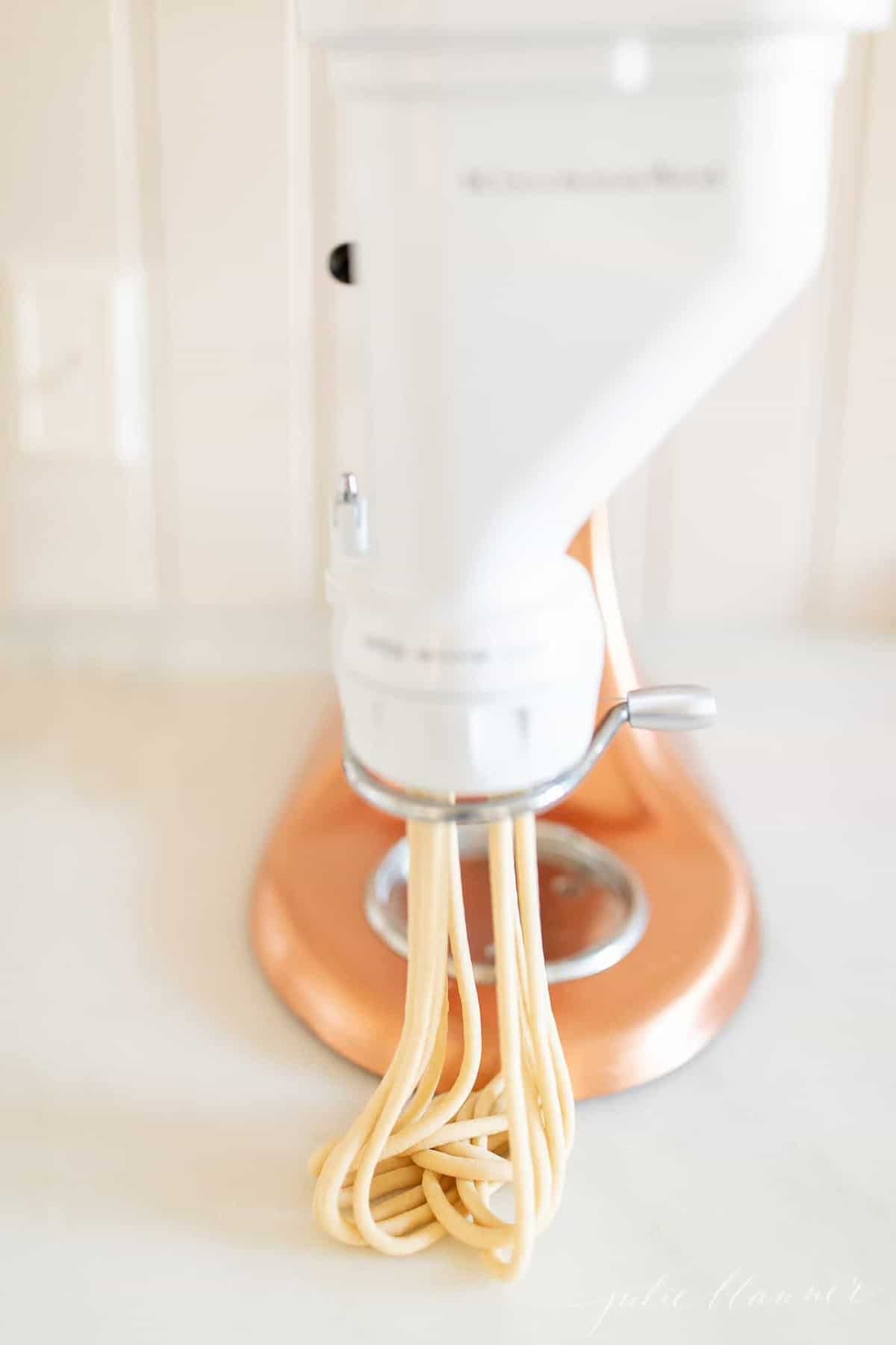 A kitchenaid stand mixer with a pasta attachment, bucatini noodles coming out