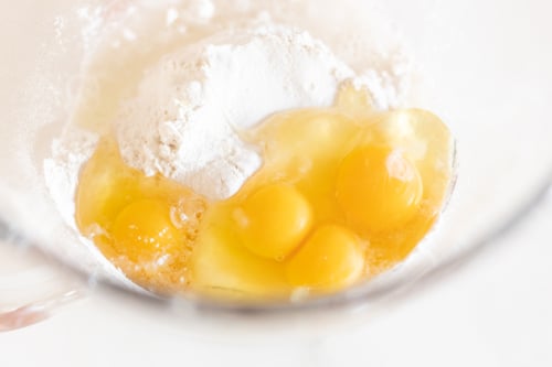 A close-up image of four raw eggs mixed with flour and bucatini pasta in a glass bowl, preparing for baking.
