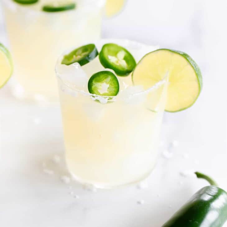 A marble surface with clear glasses full of spicy margarita recipe, jalapeno in foreground.