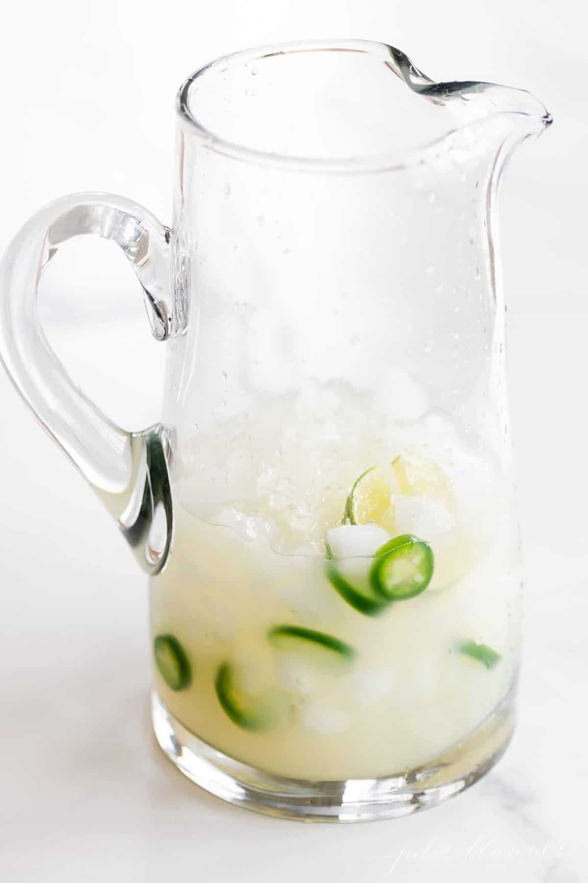 Jalapeño Margarita mix in a white pitcher, with jalapeños and limes in the mixture.