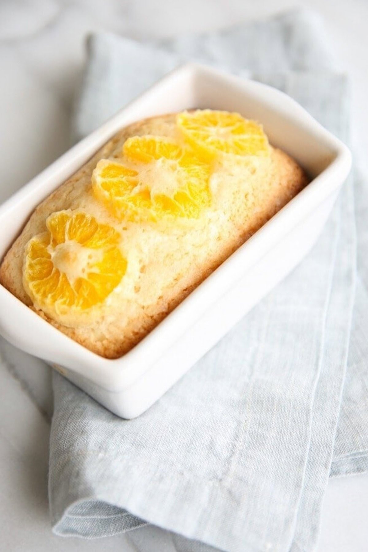 A rectangular loaf of quick orange bread, topped with three orange slices, rests in a white ceramic dish on a folded light blue cloth.