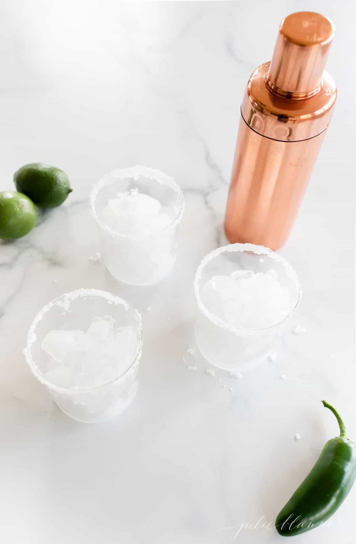 A marble surface with three glasses full of ice waiting for jalapeno margaritas.