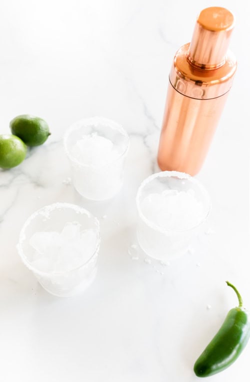 Two margaritas with salted rims, a copper cocktail shaker, limes, and a jalapeño pepper on a white marble surface for a Jalapeño Margarita.