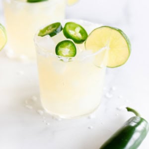 Two spicy margarita cocktails garnished with lime slices and jalapeño slices on a white surface.
