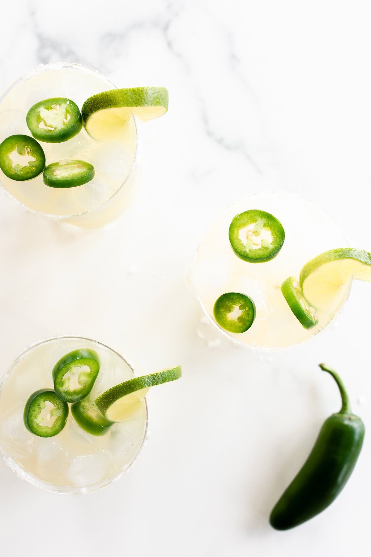 Three Jalapeño Margaritas with jalapeño slices and lime garnishes on a marble surface, with a whole jalapeño pepper beside them.