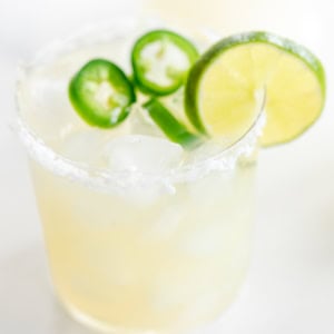 Two glasses of a spicy margarita recipe with salt rims, garnished with lime slices and jalapeño, on a white background.