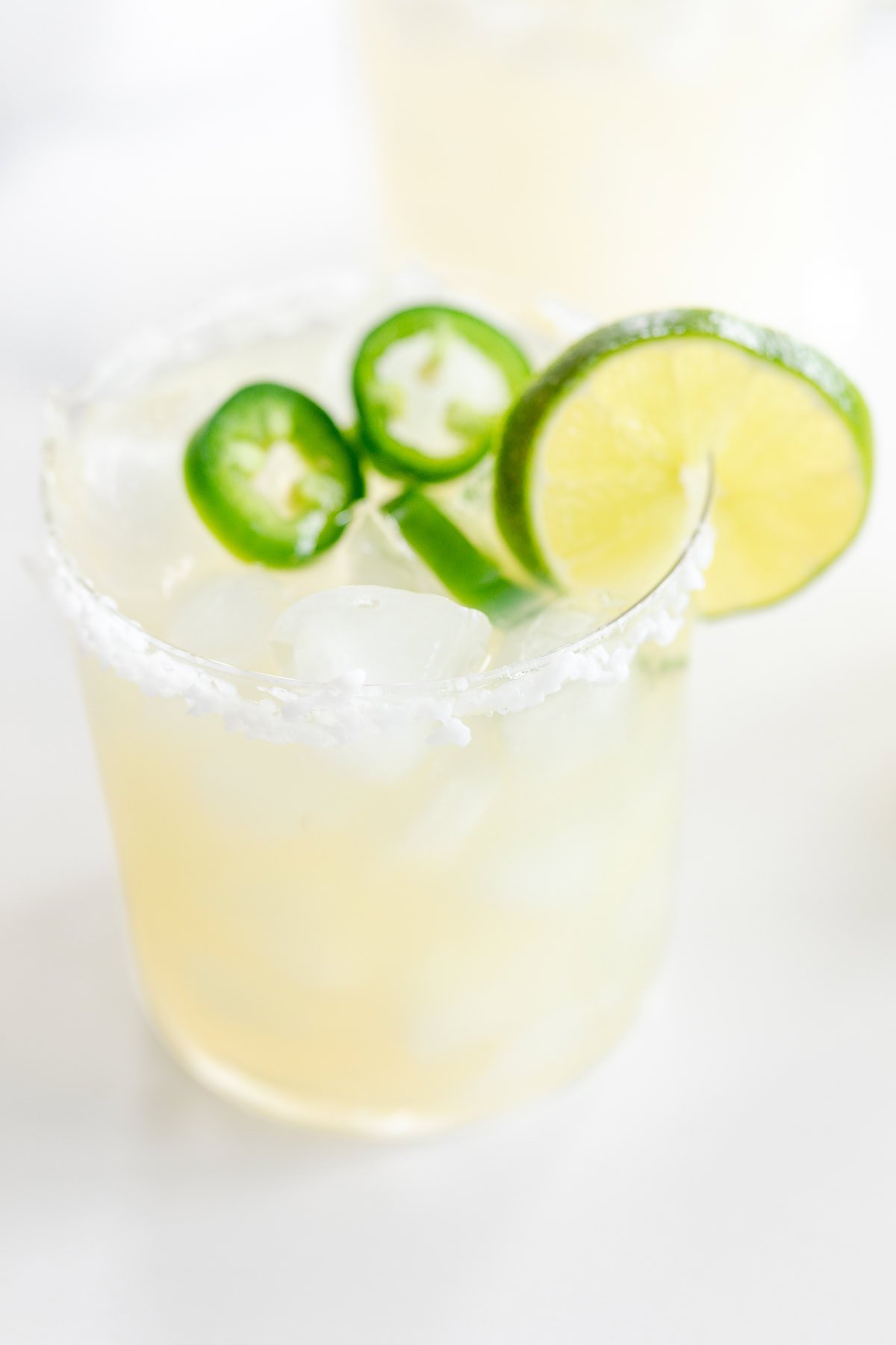 Two glasses of a spicy margarita recipe with salt rims, garnished with lime slices and jalapeño, on a white background.