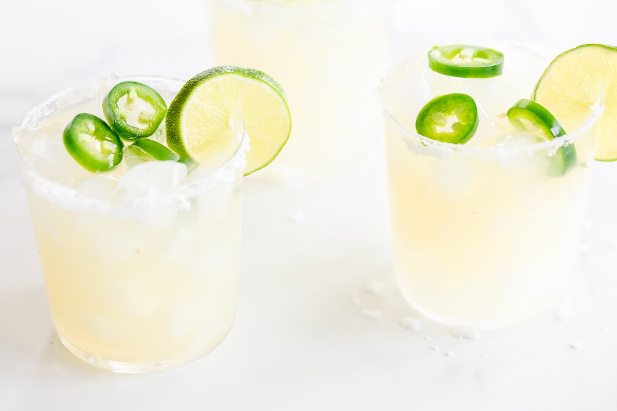 Three glasses of Jalapeño Margaritas with ice, garnished with lime slices and jalapeño rings, on a white surface.