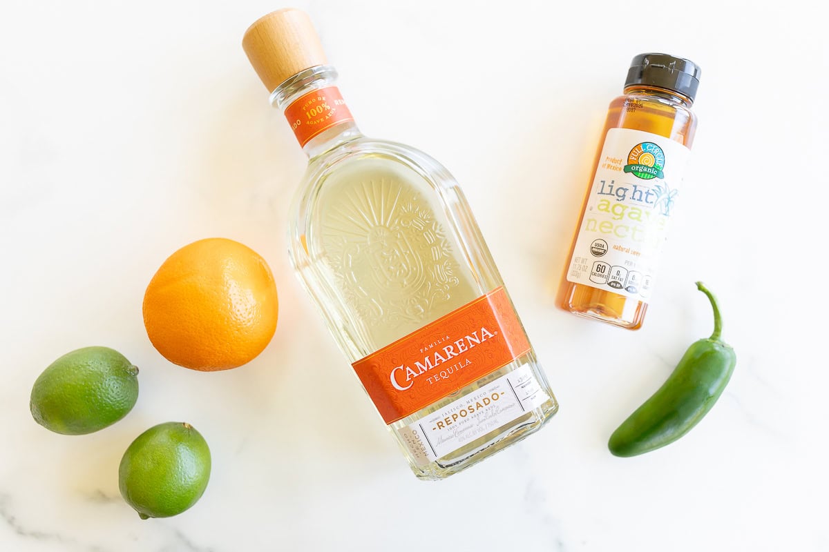 A bottle of Camarena tequila, an orange, two limes, a jar of light agave nectar, and a jalapeño pepper on a marble surface for a spicy margarita
