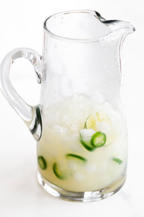 A clear glass pitcher filled with ice, sliced jalapenos and limes, containing a refreshing spicy margarita recipe.