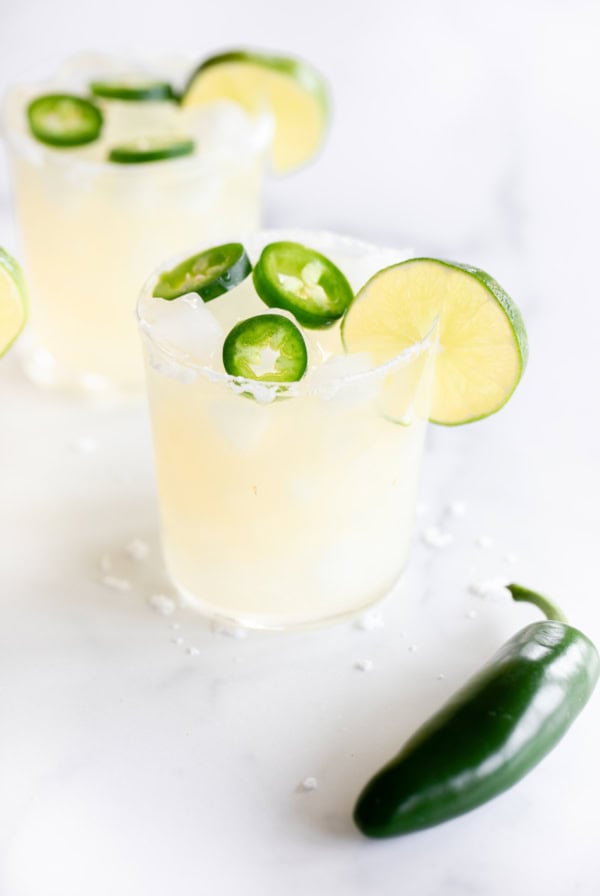 Two glasses of spicy margarita with jalapeño slices and lime garnish on a white surface, with extra jalapeño and scattered salt nearby.