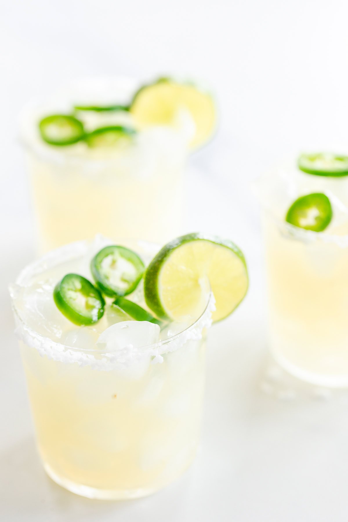 Three glasses of Jalapeño Margaritas with lime slices and ice, garnished with salt rims and jalapeño slices on a white surface.