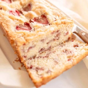 A sliced loaf of strawberry bread with a knife on a plate, delicately showcasing small pieces of strawberry baked into the crumb.